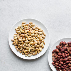Overhead view of unpeeled raw peanuts and peeled roasted peanuts on white plates, flatlay of raw and roasted peanuts on a marble countertop, process of making roasted peanuts