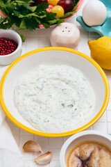 Greek yogurt sauce with lemon, garlic and parsley in a white bowl with a yellow rim on a white table with ingredients laid out around it.