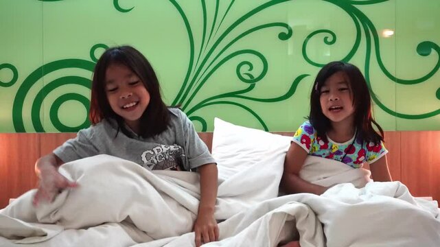 Cute little siblings hiding under blankets and playing peekaboo on a comfy bed on white pillows in the morning.