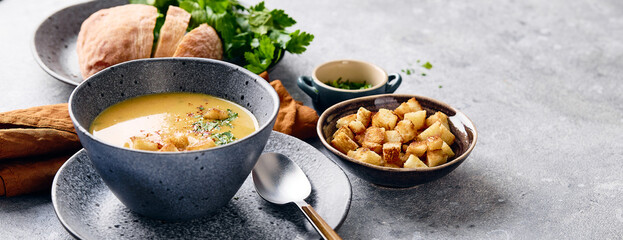 Bowl of mashed potato soup with a spoon on the plate. The bowl is filled with yellow soup puree and topped with croutons, parsley and ground red pepper.