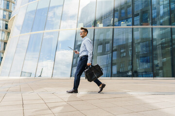 Fototapeta premium Smiling businessman in blue shirt walking with a briefcase and tablet, near modern glass buildings.