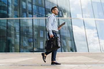 Confident businessman walking with a briefcase and tablet, dressed in smart attire, near modern glass buildings.