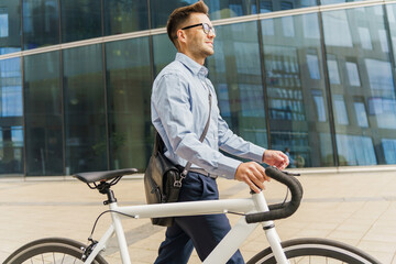 Obraz premium Smiling businessman walking his white bicycle, dressed in smart attire, beside a modern glass building.