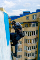 on the roof of a high-rise building against the background of multi-colored bright houses painter suspended on a safety net paints the wall blue