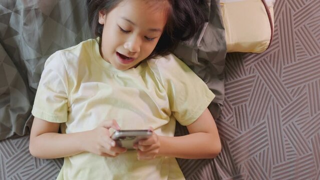 Top view of adorable Asian kid girl, lied in bed, enjoys her smartphone in a sunlit room. Her expression hints at digital fun, highlighting the allure of home tech, playing game and e-learning.