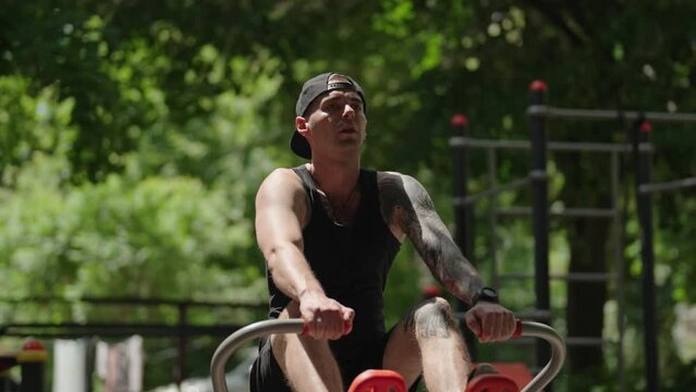 A fit young man exercising on a row machine at an outdoor gym. He is working out hard on a sunny day. The camera angle is a medium shot facing the man.