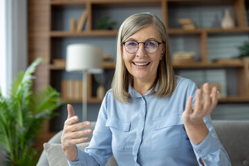 Senior woman with glasses engaging in a video call while looking and talking to the camera. She...