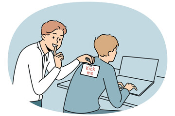 Man sticks kick me sticker on back of colleague working with laptop to make prank and laugh. Cheerful student guy jokes with friend and asks others not to talk about uplifting prank