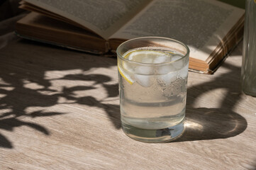 Glass with iced refreshing lemonade drink or lemon infused water, open book on wooden table background with aesthetic floral sunlight shadows, healthy summer beverage, copy space