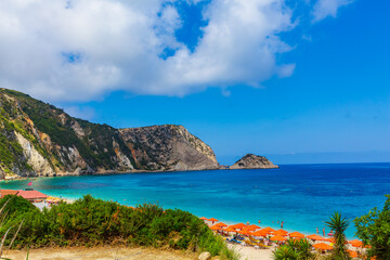 Petani Beach, Lixouri in Summer, on the beautiful Ionian island of Kefalonia, Greece, with orange umbrellas, sunbeds,  blue sky, fluffy clouds and high mountains.  Space for copy, Horizontal.