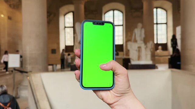 Close up of woman's hand holds phone with green screen mock up. Blurred masterpiece art at background. Concept of digital app for museums and virtual excursion.