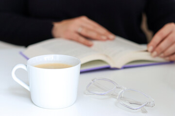Close up of a white cup with coffee and glasses on white table, blurred background with female hands and a book