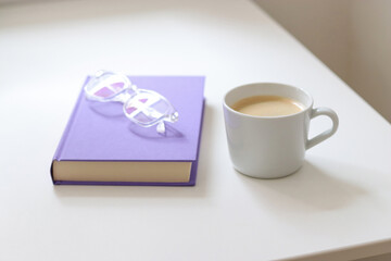 A purple mock up book with transparent glasses and a white cup of coffee on white table. Free copy space for text