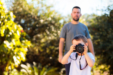 A Brazilian photographer father teaching his young son how to use a camera in a lush, green area. The father helps him frame the shot, capturing a bonding and educational moment on Father’s Day