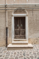 Fototapeta premium House facade with an ancient wooden door and supply lines along a small alley with nature stone pavement in a village in Sardinia, Italy