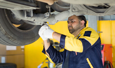 A man in uniform working underneath a car that is lifted on a hydraulic lift rack, in an automotive repair shop. The mechanic holding wrench tool securing or loosening a component under the vehicle.
