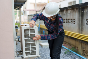 Asian female construction inspector checking air conditioning unit. Inspector wearing hard hat, ensuring proper installation and functionality, maintaining quality standards in construction site.