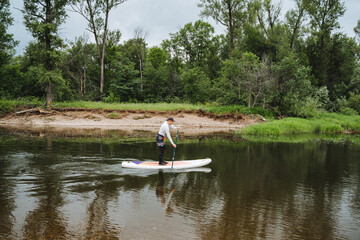 The man finds immense happiness paddle boarding in a tranquil river, surrounded by lush greenery under a blue sky, feeling a deep connection with the natural beauty surrounding him