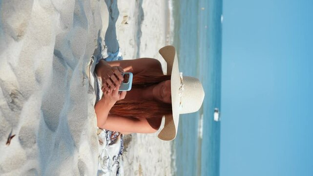 Vertical video woman sunbathes on beach tropics and chats using mobile phone. Millennial enjoy beauty of sea and beach while correspond using chat app. Using smartphone to communicate during holidays.