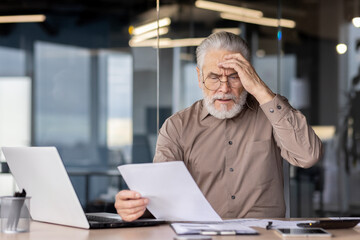 Senior businessman looking stressed while reading documents at his office desk. He is sitting in...