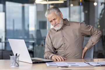 Senior man suffering from back pain while sitting at his office desk and working on a laptop. This...