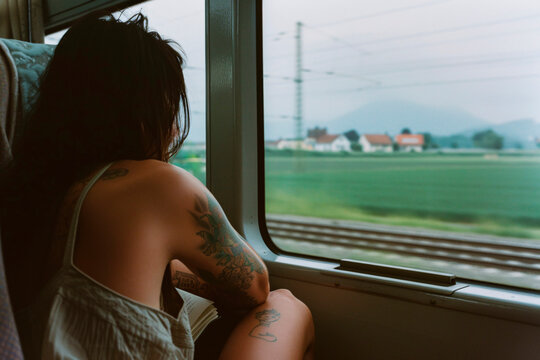 A tattooed individual is seen gazing out the window of a train, overlooking the verdant countryside, capturing a moment of deep thought and connection with nature.