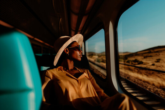 The person with a large sun hat is seen relaxing on a train, basking in the sunlight streaming through the window, capturing a moment of leisure and travel amidst beautiful scenery.