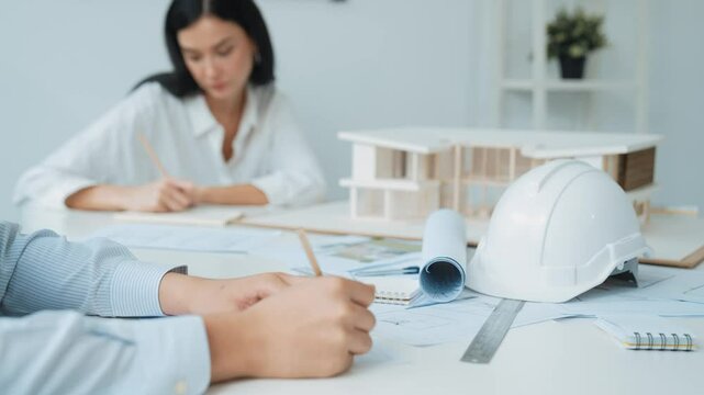 Professional architect engineer drawing blueprint and working together with young beautiful caucasian coworker on meeting table with safety helmet, house model, blueprint scatter around. Immaculate.