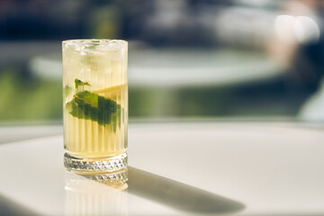 On a blurred background, a glass with lemonade or a cocktail with lemon and mint. Soft drinks or a drink with an ice cube on a light background with space to copy. High quality photo