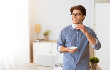 A man in a blue shirt and glasses smiles happily while enjoying a cup of coffee in his home office.