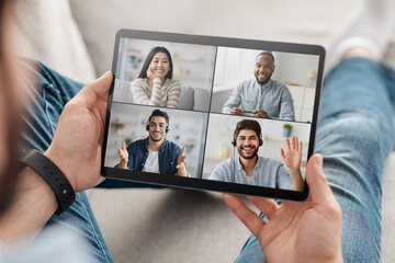 Man is holding a tablet and participating in an online video conference with four other people, relaxing on couch at home