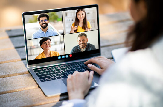 A woman sits outside on a wooden surface and participates in a video conference. Her laptop is open to a screen displaying four participants in separate video frames