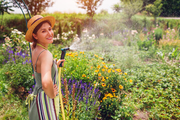 Happy woman gardener in straw hat holding garden hose watering plants in summer garden. Taking care...