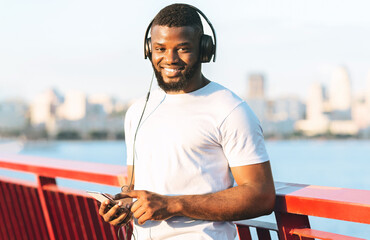 A man smiles as he listens to music on a bridge with a city skyline in the background.