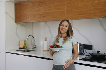 Pregnant young woman with big belly eating strawberries at the kitchen. Close up, copy space, background.
