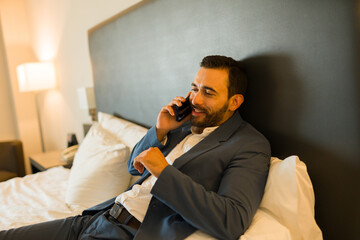 Man in business attire relaxes on a hotel room bed, chatting on the phone with a grin