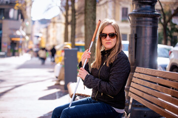 A visually impaired woman holding a white cane and sitting on a bench in the city