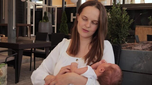 Brown-haired mother sitting comfortably in the cafe with her newborn during hot summer day looking lovingly at her child savoring the happiness and tranquility of the moment.
