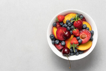 Fruit salad in a white bowl on a light background. Peach, blueberry, cherry, strawberry, plum summer mix