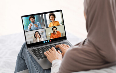 Muslim woman sits on a bed with her back to camera, using laptop to video chat with three friends. She is wearing hijab and is typing on keyboard, actively participating in online conversation.