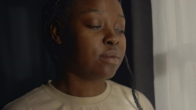 Closeup of young Black woman with ptsd looking out of window in dark room at dawn, losing contact with reality after traumatic past events