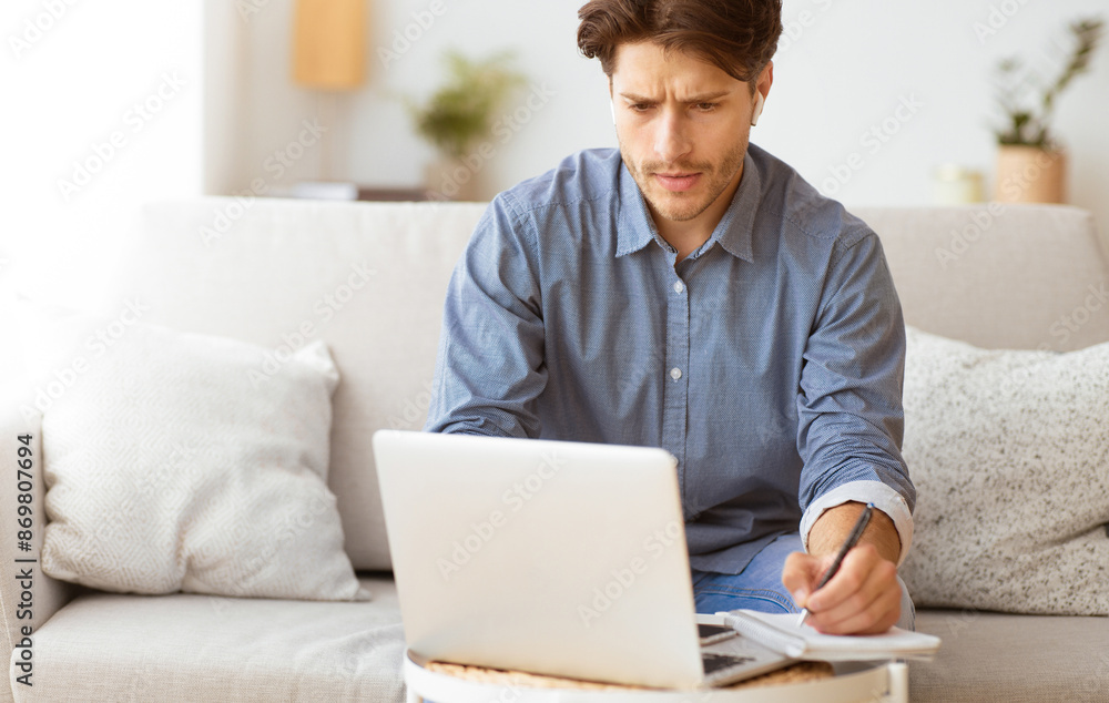 Poster A man sits on a couch, working on a laptop and taking notes in a notebook. He appears focused and engaged in his work.
