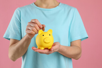 Woman putting coin into yellow piggy bank on pink background, closeup