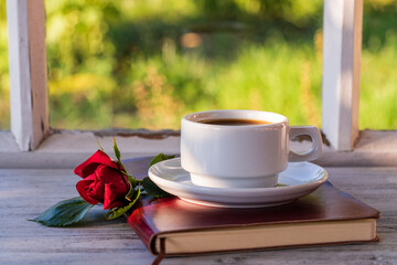 Ceramic white cup of coffee, red rose and notebook on the table in front of the window overlooking the summer garden, closeup