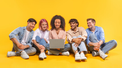 A group of five young friends are sitting on a yellow background, looking at a laptop screen together. They are all smiling and appear to be enjoying each others company.