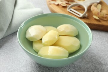 Fresh raw potatoes in bowl, peeler and peels on light grey table