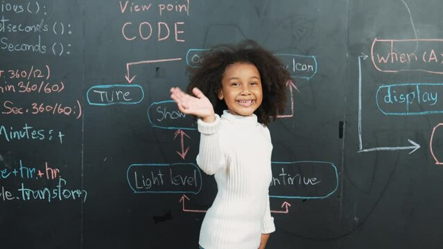 African girl smiling and waving at camera while standing at board with engineering prompt or coding, programing system written in STEM classroom. Child looking at camera while greeting. Erudition.