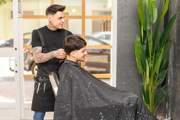 tattooed barber cutting the hair and beard of a young, dark-haired client.
