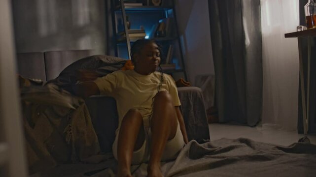 Full handheld shot of young African American woman shouting at phone during call and undergoing anger attack being triggered by heard words, throwing things in rage while sitting on floor by bed
