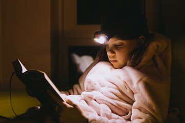 Child is engrossed in reading a book at night, illuminated by a headlamp.